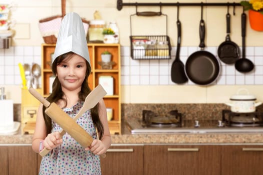 Happy asian little girl with chef hat holding cooking appliance in the kitchen room