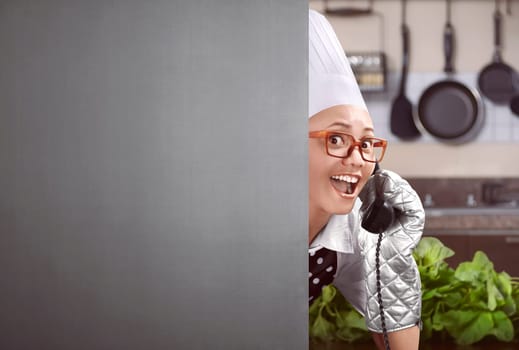 Happy asian female chef lifted up the phone from behind grey banner on the kitchen