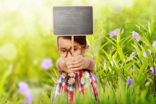 Happy asian little girl holding blank blackboard over green garden background