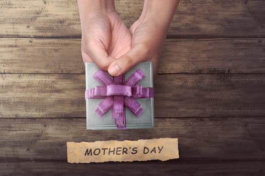 People hand holding small gift with Mother's Day greeting on wooden table