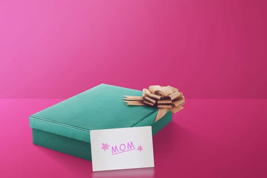 Gift box with gold ribbon and mom text on paper over pink background