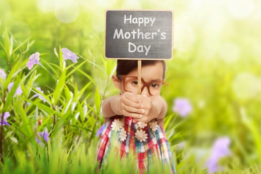 Funny asian child holding chalkboard with Happy mothers day message over green garden background