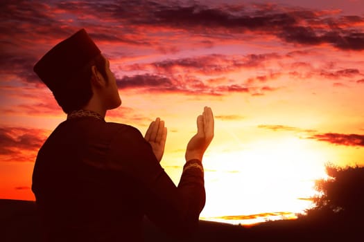 Back view of asian muslim man with black cap praying against sunset background