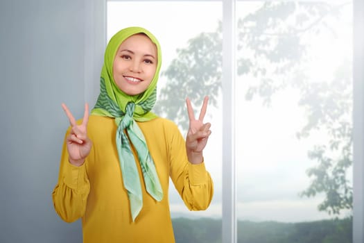 Young asian woman muslim giving peace sign against indoor background