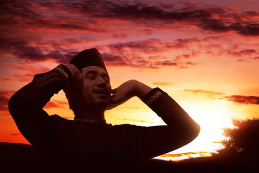Young asian muslim man praying with raising hand at sunset background