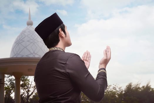 Back view of asian muslim man raising both his hand to pray with blue sky background