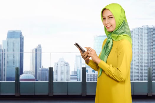 Smiling asian muslim woman using a smartphone against skyscraper background