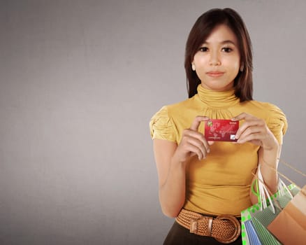 Happy asian woman with many shopping bags and credit card in her hand against grey background