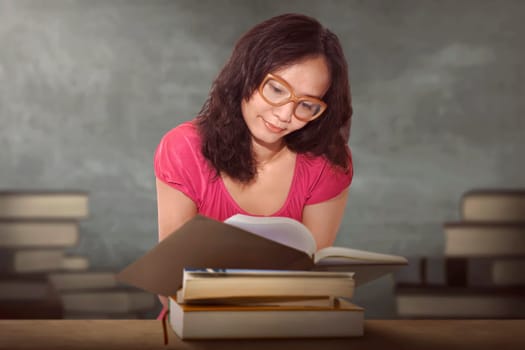 Charming asian woman sitting by wooden table and reading book with stacked book on background