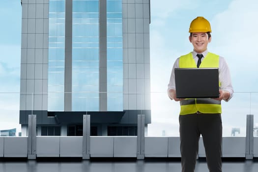 Handsome asian architect man with laptop wearing protective helmet standing in front of a skyscrapers building
