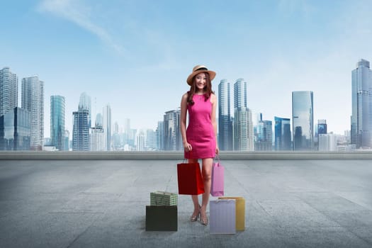 Young asian woman wearing hat holding shopping bags against cityscape background