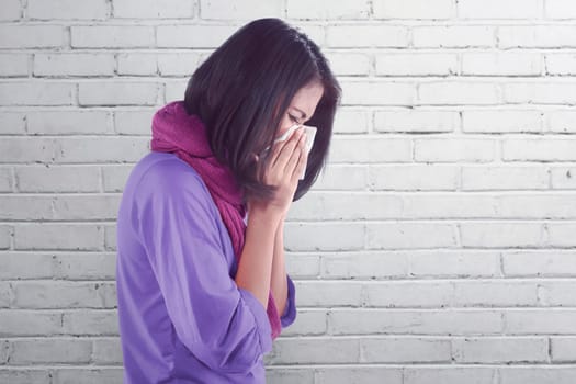 Sick asian woman with flu and fever holding tissue in nose against wall background