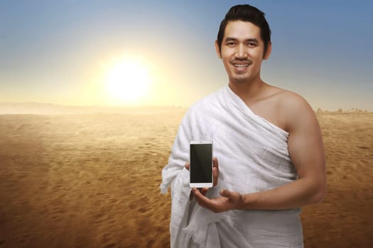 Handsome asian muslim man with ihram clothes showing phone against sunset background