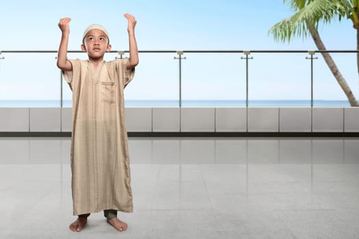 Asian muslim kid with cap raising hand and praying on terrace mosque