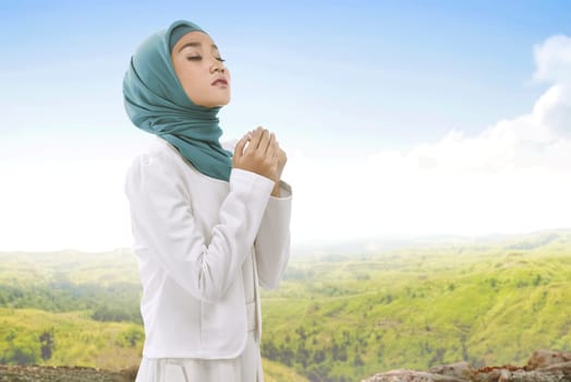 Pretty asian muslim woman wearing hijab raising hand and praying with landscape view background