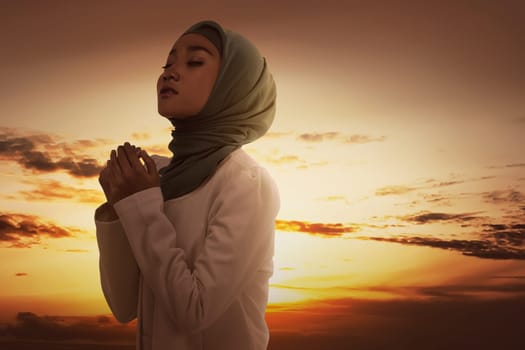 Beautiful asian muslim woman in veil praying to god during sunset at sky