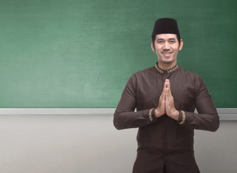 Cheerful asian muslim man wearing traditional dress with hand gesture against chalkboard background for copyspace