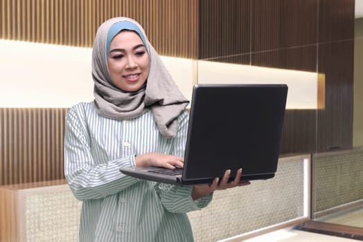 Beautiful asian muslim woman standing while typing on laptop inside her office room