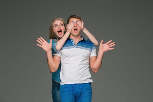 Portrait of a young surprised couple standing against gray studio background. The girl closing eyes to the guy