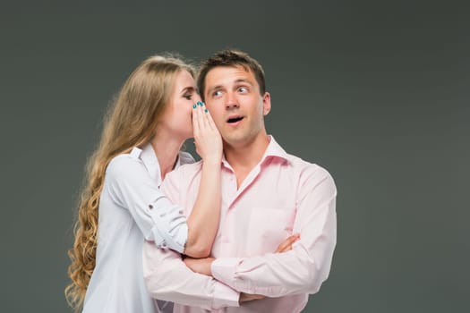 Portrait of a young couple standing against gray studio background. woman whispering in man ear