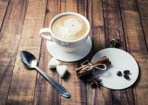 Fresh tasty coffee with spices. Coffee cup, cinnamon sticks, coffee beans, anise, sugar, spoon and coasters on vintage wooden kitchen table background.
