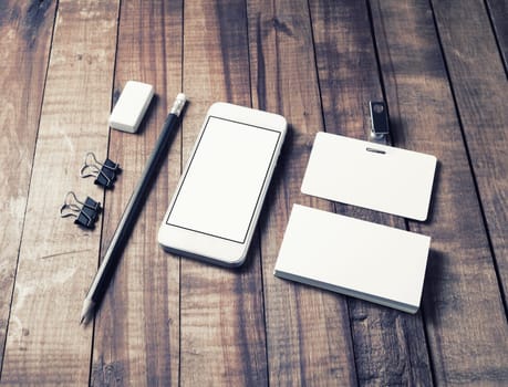 Photo of smartphone with blank white screen, business cards, badge, pencil, eraser and metal clamps on wooden table background. Blank stationery set.