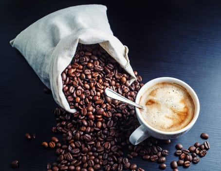 Coffee cup and coffee beans scattered from a canvas bag on black background.