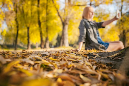 Pretty blonde sitting in a wooden old boat which is on the ground laid by yellow fallen autumn leaves. dressed gray coat, a dress and tights.