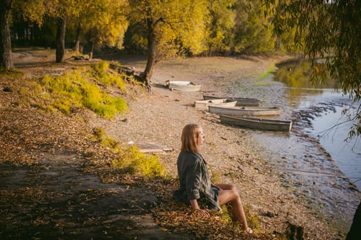 pretty blonde girl in sneakers, nylon stockings and coat, sits on  banks of river, on yellow leaves, enjoying warmth of autumn sun rays