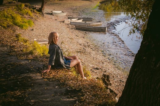 pretty blonde girl in sneakers, nylon stockings and coat, sits on  banks of river, on yellow leaves, enjoying warmth of autumn sun rays