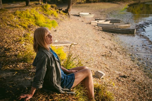 pretty blonde girl in sneakers, nylon stockings and coat, sits on  banks of river, on yellow leaves, enjoying warmth of autumn sun rays