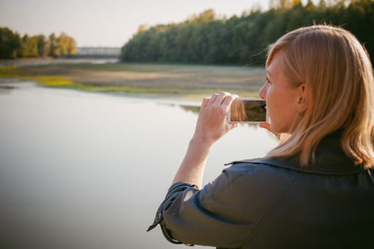 pretty blonde girl in sneakers, nylon stockings and coat. walks and photographed on a smartphone selfi on the stone shore of the lake in the last sunshine of the departing summer.