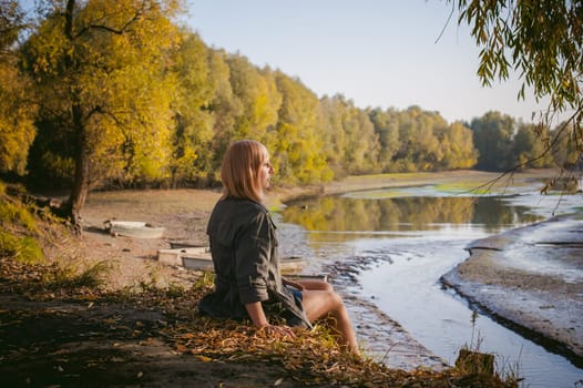 pretty blonde girl in sneakers, nylon stockings and coat, sits on  banks of river, on yellow leaves, enjoying warmth of autumn sun rays