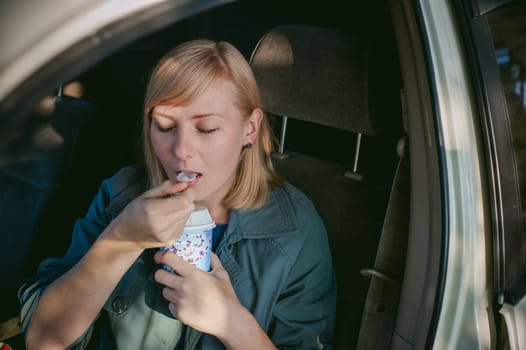 girl eating ice cream with a spoon in the car. blond girl in a raincoat, eats ice cream while in the car during a road trip