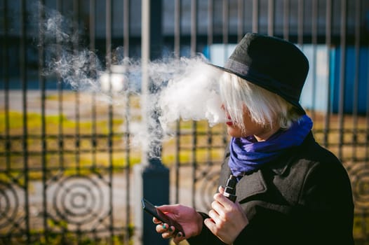 portrait of young beautiful woman with white hair, in a black coat, a skirt and a black hat, smoking an electronic cigarette, blowing the smoke vapor