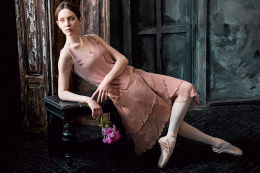 Young and incredibly beautiful ballerina is posing in a black wooden studio with flowers