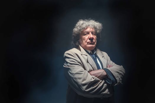 The senior man in cloak standing as detective or mafia boss. Studio shot on gray in retro stile in the light and smoke ray
