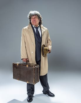 The senior man in cloak as detective or mafia boss. Studio shot on gray in retro stile. Mature man with hat and suitcase