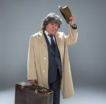 The senior man in cloak as detective or mafia boss. Studio shot on gray in retro stile. Mature man with hat and suitcase