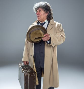 The senior man in cloak as detective or mafia boss. Studio shot on gray in retro stile. Mature man with hat and suitcase