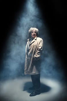 The senior man in cloak standing as detective or mafia boss. Studio shot on gray in retro stile in the light and smoke ray
