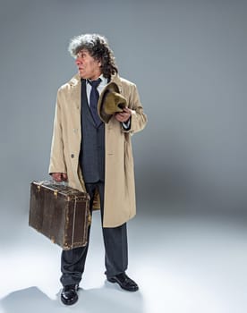 The senior man in cloak as detective or mafia boss. Studio shot on gray in retro stile. Mature man with hat and suitcase