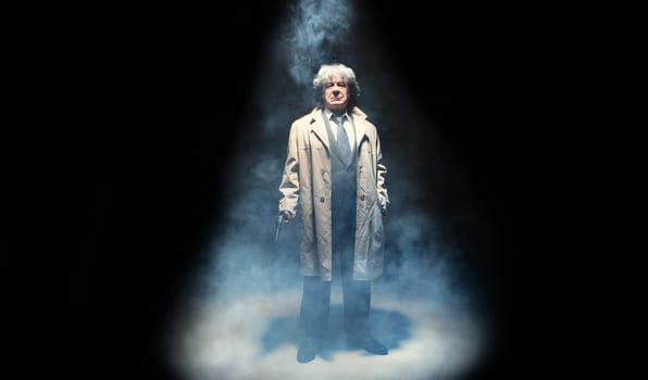 Senior police agent with a gun standing as detective or mafia boss. Studio shot on gray in retro stile in the light and smoke ray