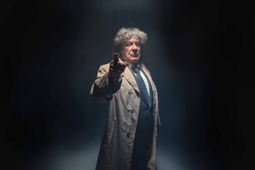 The senior man in cloak standing as detective or mafia boss and pointing at camera. Studio shot on gray in retro stile in the light and smoke ray