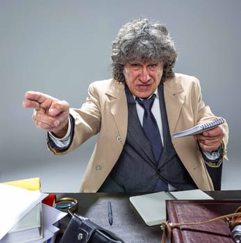 The senior man in cloak as detective or mafia boss pointing at camera at table. Studio shot on gray in retro stile