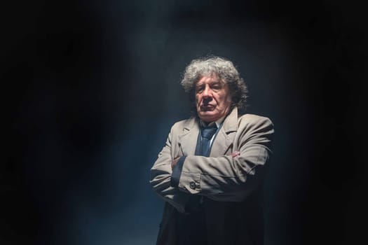 The senior man in cloak standing as detective or mafia boss. Studio shot on gray in retro stile in the light and smoke ray