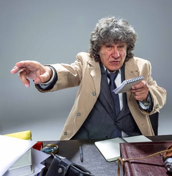The senior man in cloak as detective or mafia boss pointing at camera at table. Studio shot on gray in retro stile