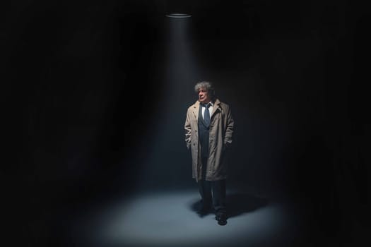 The senior man in cloak standing as detective or mafia boss. Studio shot on gray in retro stile in the light and smoke ray