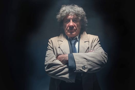 The senior man in cloak standing as detective or mafia boss. Studio shot on gray in retro stile in the light and smoke ray