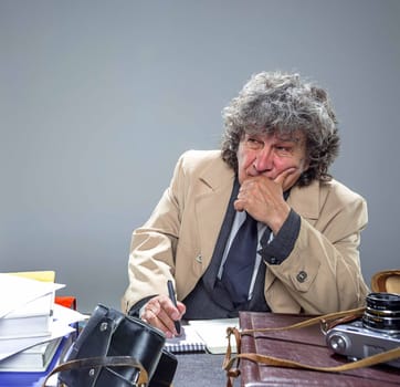 The senior man in cloak as detective or mafia boss writing in a notebook with pen at table. Studio shot on gray in retro stile